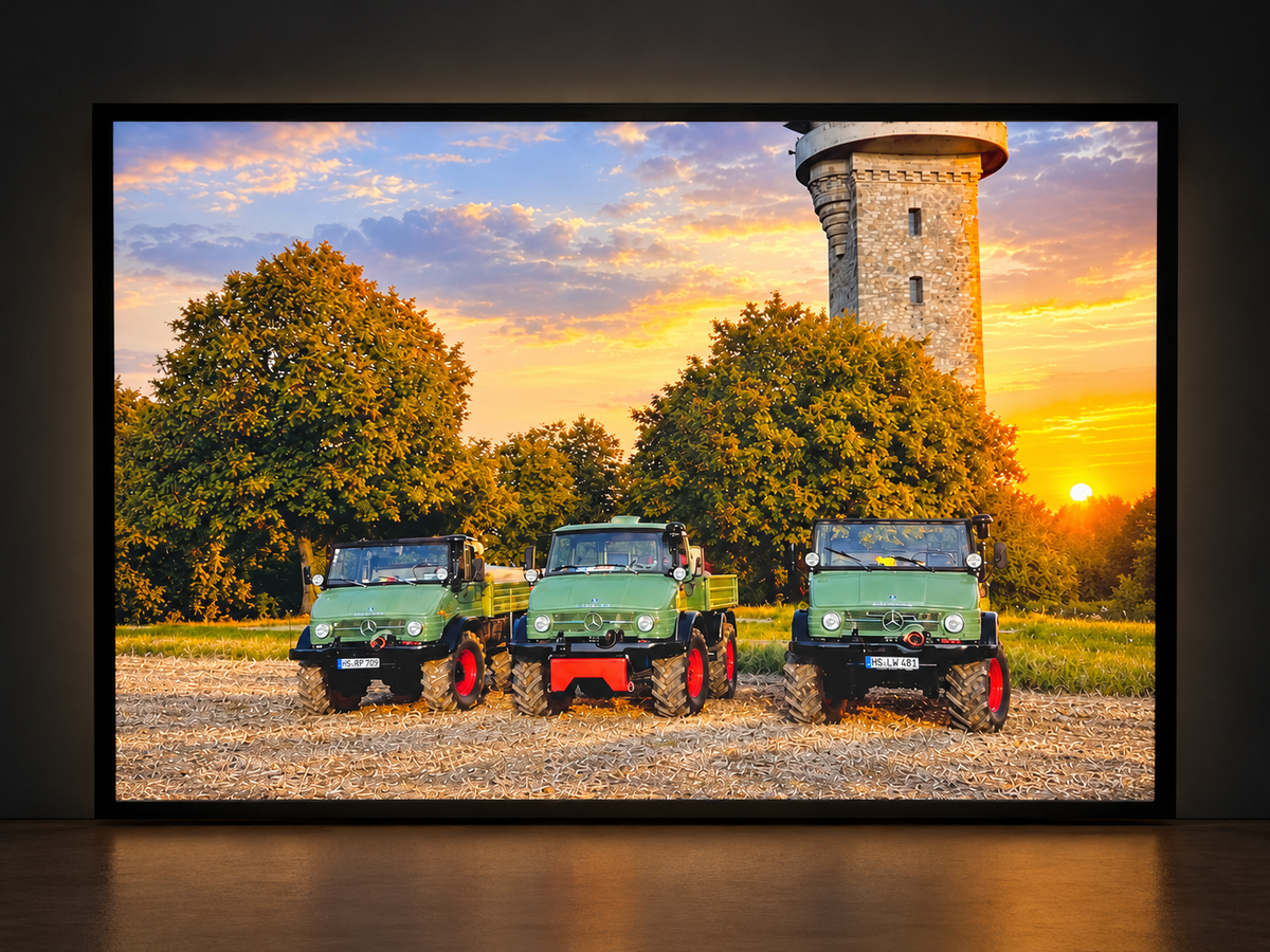 Illuminated photo lightbox featuring three green Unimog vehicles beneath a golden sunset